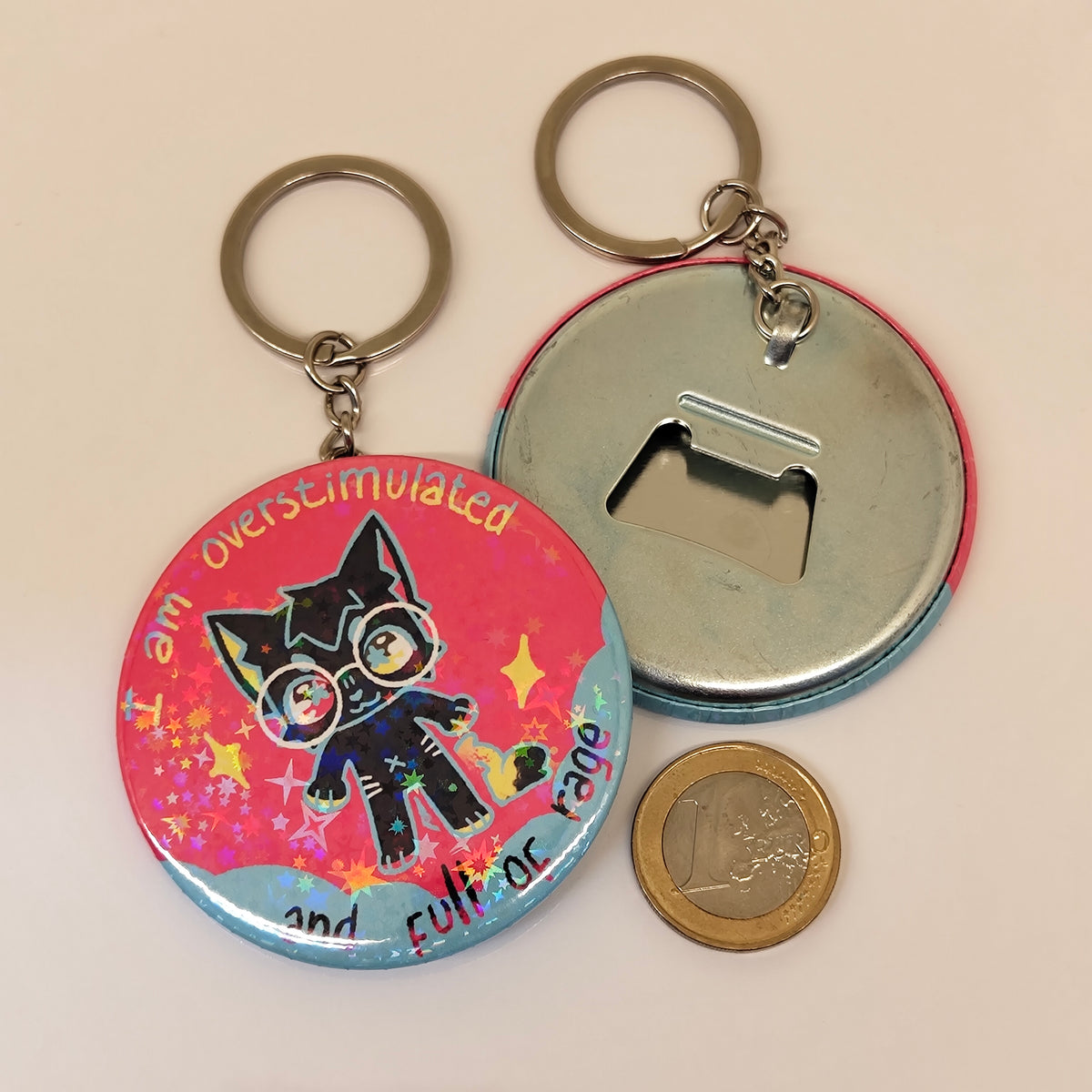 A round metal bottle opener keychain shown from both sides on a plain light background. One side features a colorful graphic of a black cartoon cat with glasses on a pink and blue background with the text 'I am overstimulated and full of rage'; the other side shows the plain metal bottle opener back with a key ring.